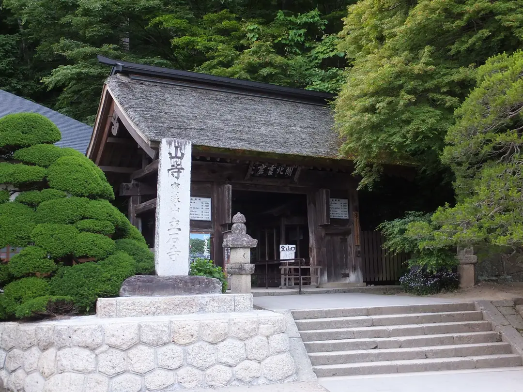立石寺參拜道入口的茅葺屋頂木門，門前立著寫有山寺寶珠山立石寺的石碑與石燈籠，右邊是通往門內的石階