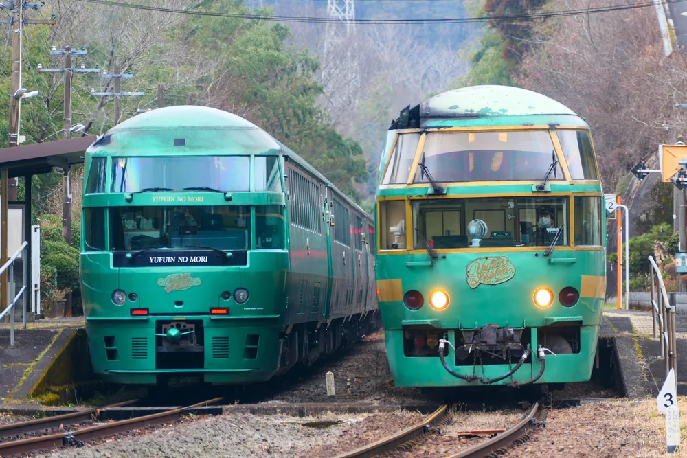 兩列綠色的 JR 九州特急ゆふいんの森列車在久大本線的小站並排停靠，左邊是新款、右邊是舊款車頭