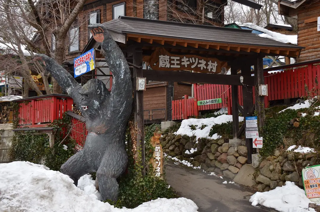 殘雪中的蔵王キツネ村 入口，木造門樓上掛著寫有蔵王キツネ村 的木匾，兩側是紅色柵欄與石牆